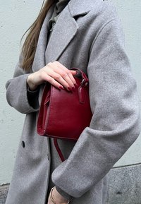 Borsa a tracolla rossa in pelle con design strutturato, texture liscia e manico piccolo. Indossata sotto un cappotto grigio di lana con maniche ampie.
