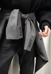 Tissu gris noué autour de la taille, présentant un motif texturé et des boutons, associé à un haut noir et un jean foncé.