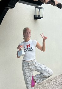 White fitted t-shirt featuring red text and a bow graphic, paired with gray snake-print wide-leg jeans, and pink clogs, while holding a clear cup. Set outdoors.