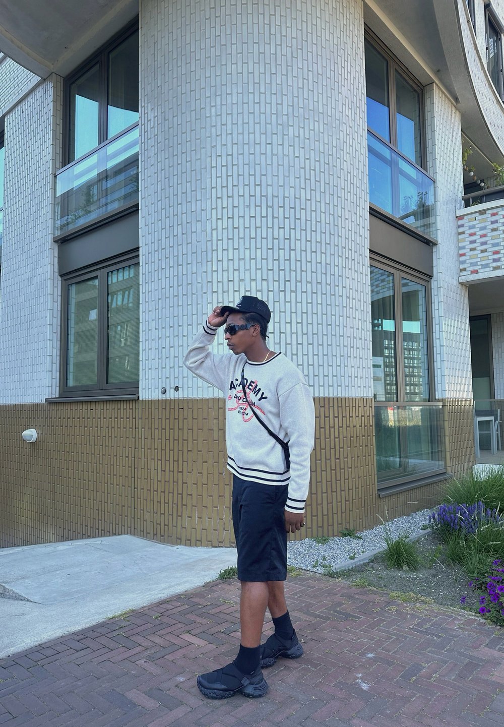 Outfit casual composto da una felpa bianca con dettagli neri, pantaloni corti neri e sneakers nere. In piedi vicino a un muro di mattoni testurizzati.