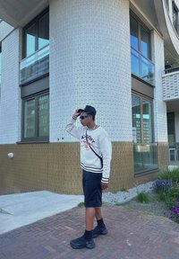 Outfit casual composto da una felpa bianca con dettagli neri, pantaloni corti neri e sneakers nere. In piedi vicino a un muro di mattoni testurizzati.