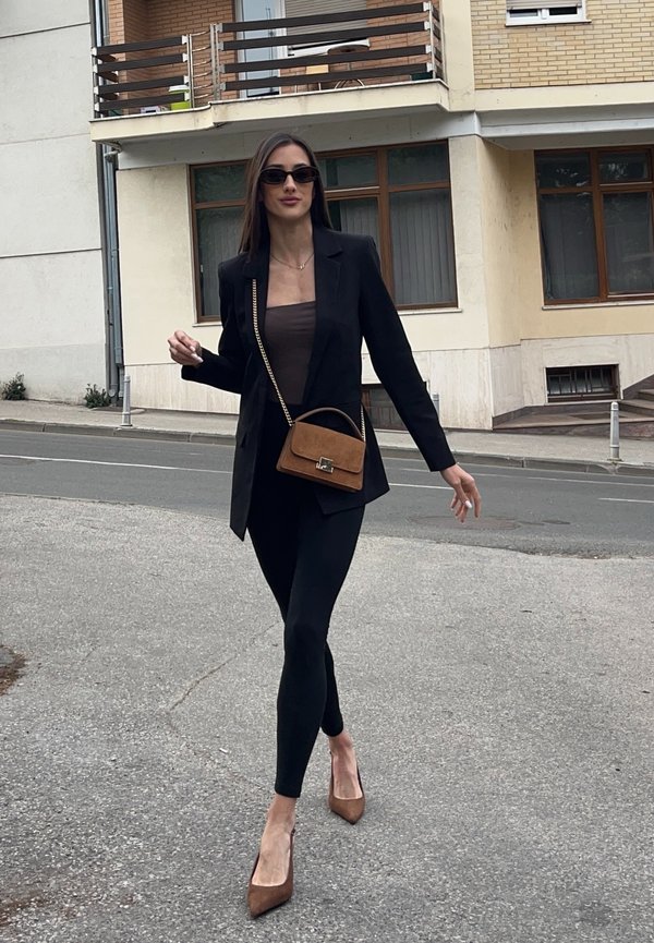Femme marchant dans la rue portant un blazer noir, des leggings, un haut marron, des talons marron, des lunettes de soleil et un petit sac à main marron avec une chaîne dorée.