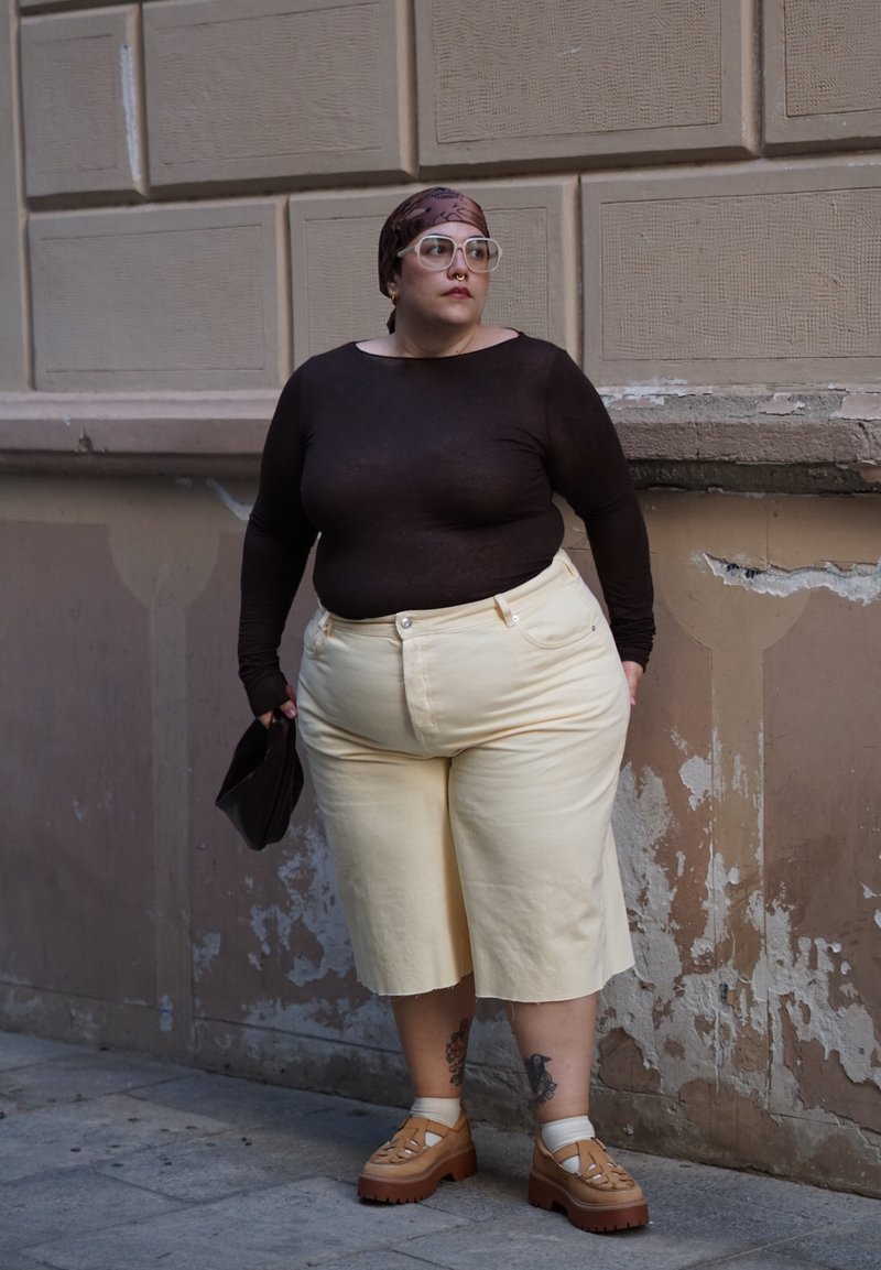 Personne avec des lunettes et un foulard sur la tête portant un haut noir, un pantalon crème et des chaussures marron, se tenant contre un mur usé tenant un sac noir.