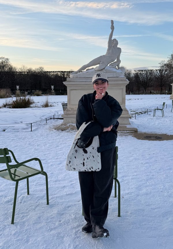 Personne en vêtements d'hiver et casquette, debout en souriant avec une main sur le menton, grand sac tacheté sur le bras, sol couvert de neige, et statue allongée en arrière-plan.