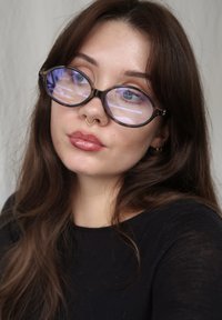 Gafas de montura redonda y negra con lentes tintados en azul. El modelo tiene un aro septal dorado, labios brillantes y cabello largo y castaño. Lleva una blusa negra. Fondo neutro.