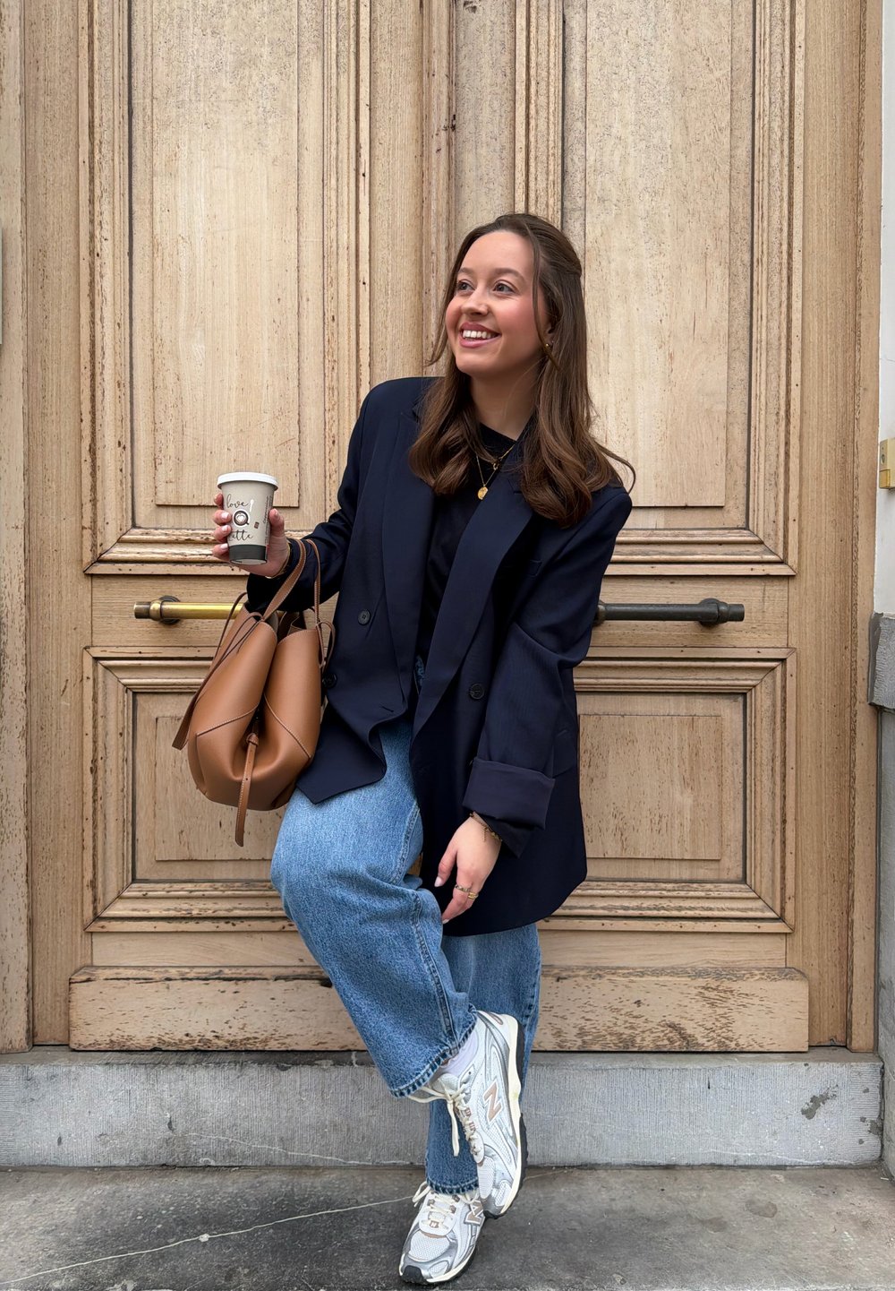 Jeune femme en blazer marine et jean tenant une tasse de café et un sac à main marron, souriant en posant devant une grande porte en bois.