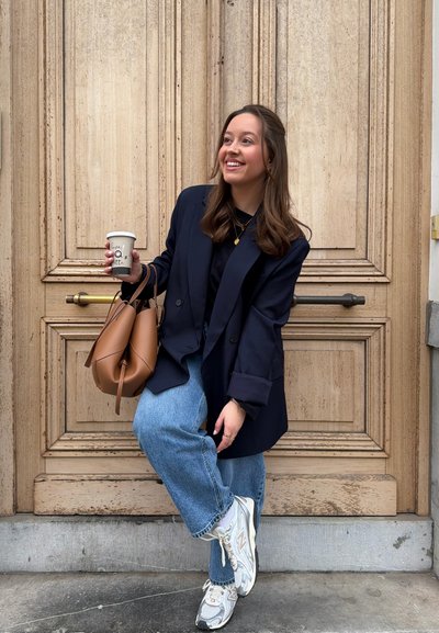 Joven mujer con blazer azul marino y jeans sosteniendo una taza de café y un bolso marrón, sonriendo mientras posa frente a una gran puerta de madera.