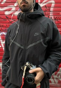 Homme en veste noire Nike tenant un appareil photo Canon vintage, debout devant une porte à volet métallique rouge couverte de graffitis.