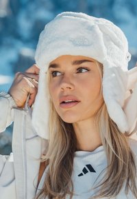 Biela klobúk s umelým kožúškom a klapkami na uši, s logom The North Face. Mäkká textúra, ľahký dizajn, nosený s prešívanou bundou.