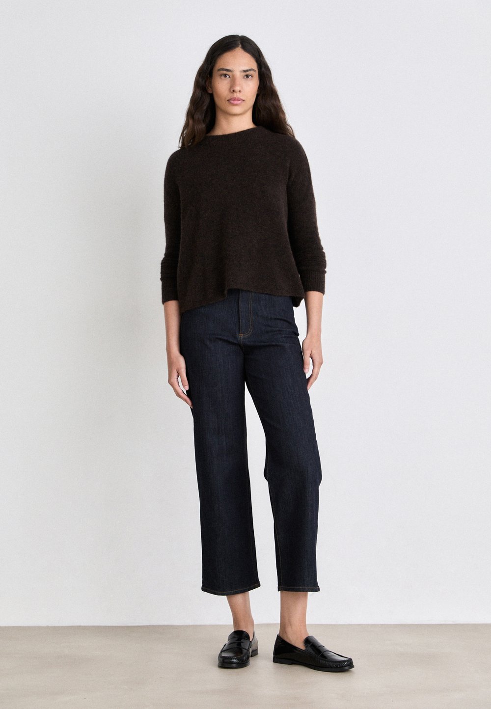 Maglione marrone con texture e scollatura ampia, abbinato a jeans blu scuro alla caviglia e mocassini neri, su uno sfondo chiaro.