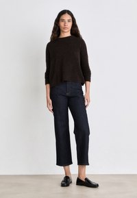 Maglione marrone con texture e scollatura ampia, abbinato a jeans blu scuro alla caviglia e mocassini neri, su uno sfondo chiaro.