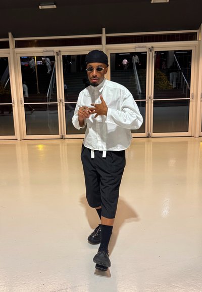 Camisa blanca de manga larga con un alto cuello, pantalones cortos a rayas negras y zapatos negros. Los accesorios incluyen gafas de sol redondas y un gorro negro.