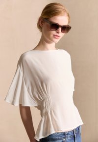 Top bianco leggero con maniche corte e lati arricciati, abbinato a jeans blu. Il modello indossa anche occhiali da sole scuri, rettangolari.