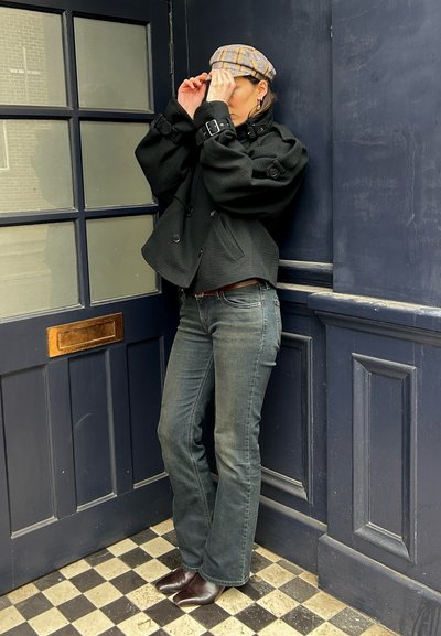 Persona ajustándose una gorra plana a cuadros, vistiendo una chaqueta negra corta, vaqueros grises y botas marrones puntiagudas, de pie en una esquina azul oscuro junto a una puerta.