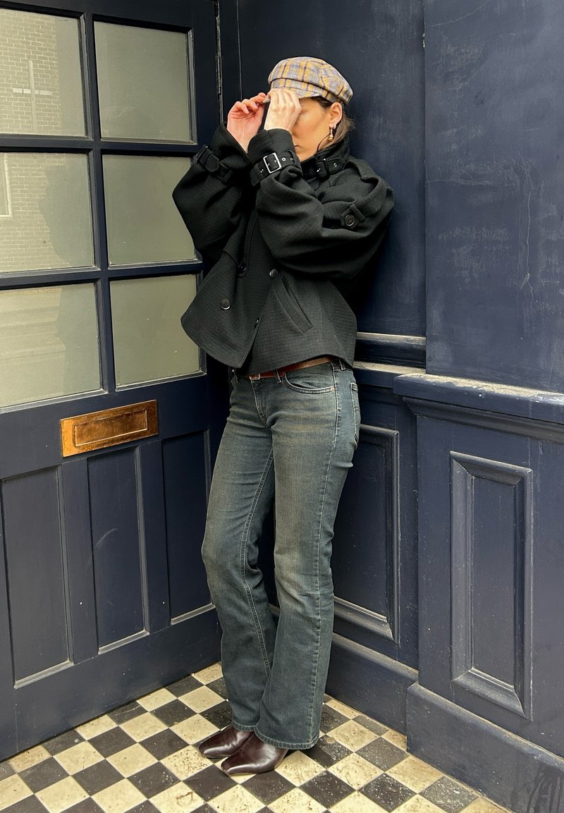 Persona che sistema un berretto piatto a quadri, indossa una giacca nera corta, jeans grigi e stivali marroni appuntiti, in piedi in un angolo blu scuro vicino a una porta.