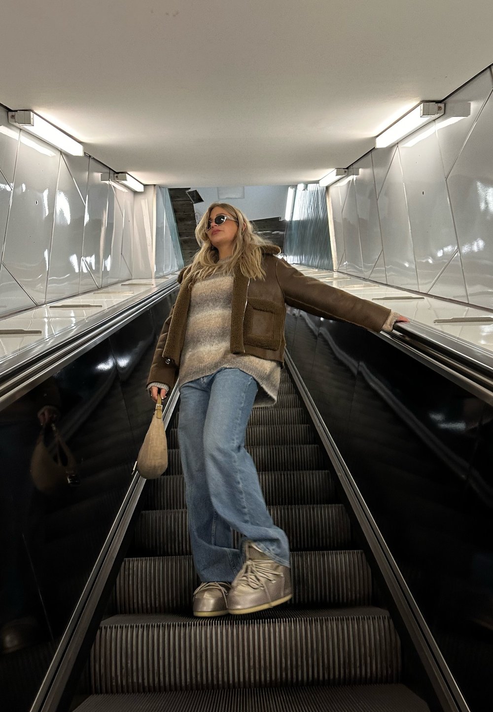 Smeđa jakna od ovčje kože s podstavom od krzna preko prugastog pulovera, svjetloplave široke traperice i srebrne tenisice, drži bež torbu na pokretnim stepenicama.