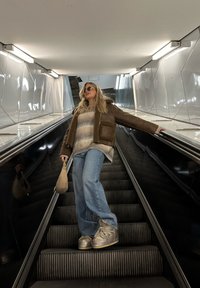 Smeđa jakna od ovčje kože s podstavom od krzna preko prugastog pulovera, svjetloplave široke traperice i srebrne tenisice, drži bež torbu na pokretnim stepenicama.