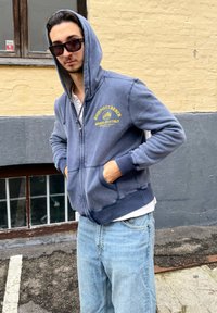 Felpa con zip di colore blu navy, dotata di tasca frontale e polsini a costine, con testo giallo. Indossata sopra una maglietta bianca e jeans azzurri.