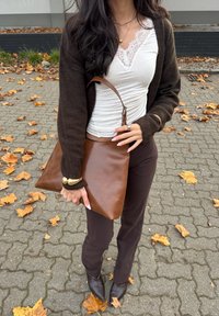Brown leather handbag with a triangular shape, paired with a brown cardigan, white top, and fitted brown trousers. Ground covered in fallen leaves.