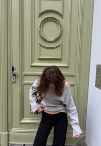 Porta in legno verde chiaro con disegni in rilievo intricati e accenti circolari. Maglione grigio a spalle scoperte, pantaloni neri, con una borsa decorata.