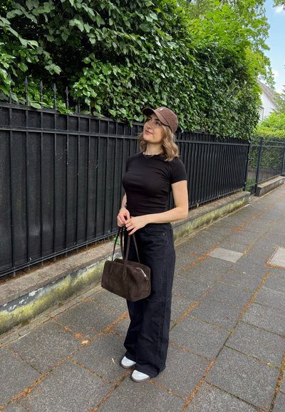 Camiseta negra ajustada, jeans negros de pierna ancha de cintura alta, gorra marrón y un bolso de ante marrón oscuro. Deportivos grises con suelas blancas. Follaje verde de fondo.