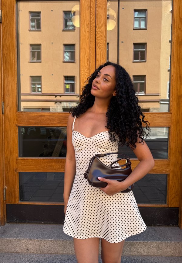 Femme aux cheveux bouclés portant une robe blanche à pois tient un sac à main noir, debout devant des portes vitrées encadrées de bois.