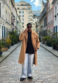 Beige kappa, svart skjorta och ljusblå vida jeans; urban gata med gatstenar och krukväxter i bakgrunden.