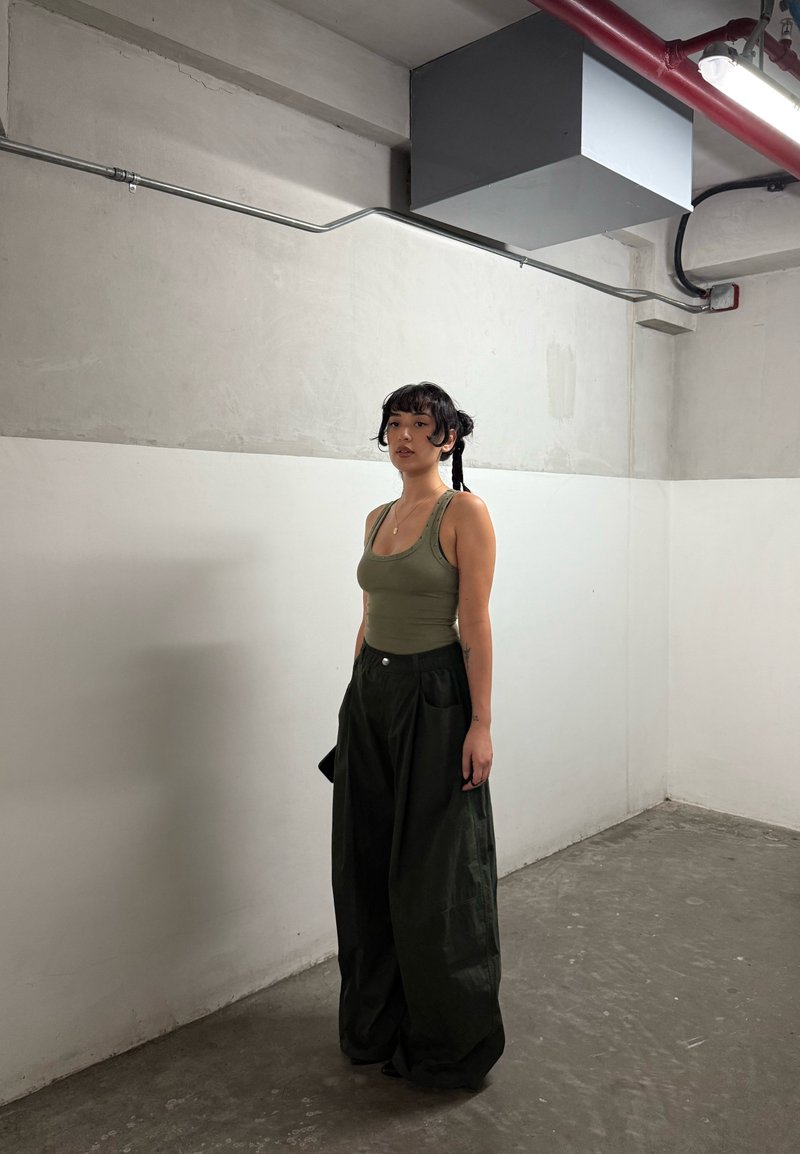 Jovem mulher com cabelo trançado, vestindo uma camiseta sem mangas verde-oliva e calças largas pretas, em pé numa sala industrial com paredes e piso de betão.