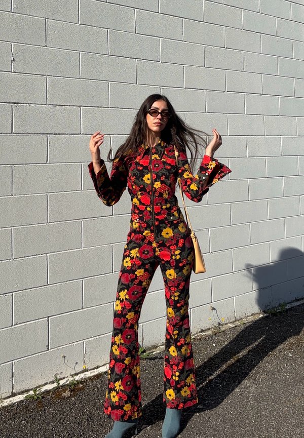 Kvinde iført en sort jumpsuit med rødt og gult blomsterprint, solbriller, holder sit hår, står op ad en lys grå murstensvæg i sollys.