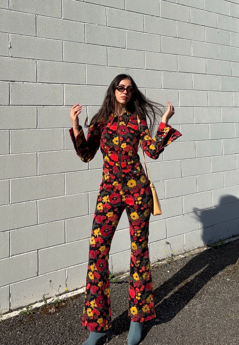 Femme portant une combinaison noire à imprimé floral rouge et jaune, lunettes de soleil, tenant ses cheveux, debout contre un mur de briques gris clair au soleil.