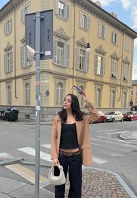 Béžový blazer cez čierny top, modré džínsy, drží slnečné okuliare a svetlú tkanú tašku. Mestskej prostredie s budovou a cestnými značkami.