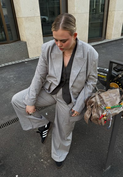 Traje gris texturizado de corte holgado, combinado con una camisa oscura abotonada. Zapatillas negras y un bolso beige con llaveros coloridos.