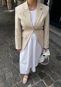 Μπεζ κομψό σακάκι με κούμπωμα, λευκή ριμπ τοπ και ροή λευκού φορέματος; συνδυασμένο με διαφανή σανδάλια με λουρί και μικρή λευκή τσάντα.