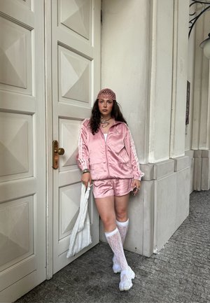 Chándal deportivo de satén rosa con rayas blancas, combinado con calcetas blancas hasta la rodilla y zapatillas blancas. Un gorro a juego completa el look.