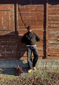 Jeune homme portant un bonnet noir, des lunettes de soleil, un pull sombre et un jean, appuyé contre un mur en bois patiné, avec un grand sac posé au sol à proximité.