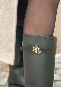 Juodos kelio ilgio lietaus botai su auksiniu „RL“ ženklu, dėvimi ant skaidrių juodų pėdkelnių, derinami su smėlio spalvos vilnoniu paltu.