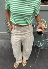 Grün-weißer gestreifter Crop-Top, beigefarbene weit geschnittene Hose mit Taschen und gelbe Flip-Flops. Hält eine kleine schwarze Kaffeetasse.