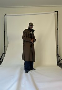 Mann in braunem Trenchcoat, Camouflage-Cap, Brille, dunklen weit geschnittenen Hosen und schwarzen Schuhen steht im Studio vor einem weißen Hintergrund.