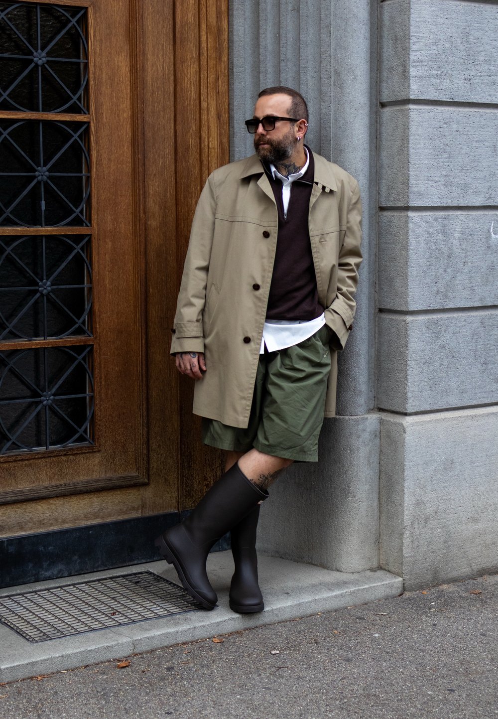 Trench beige sur un pull sombre, une chemise blanche et un short vert. Bottes en caoutchouc noires. Adossé à un mur en pierre texturé.