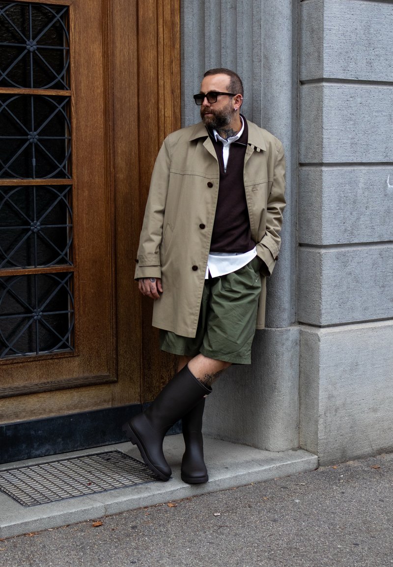 Beige trench coat over a dark sweater, white shirt, and green shorts. Black rubber boots. Leaning against a textured stone wall.