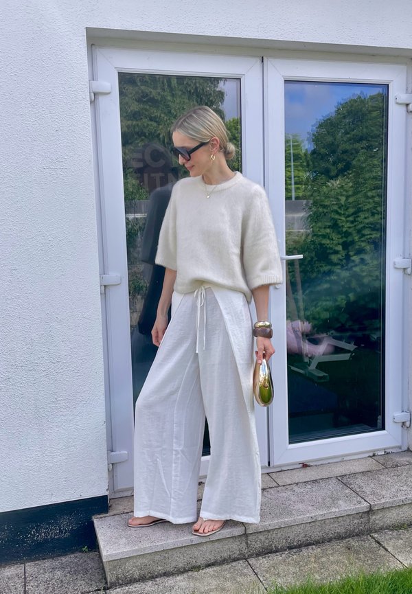 Vrouw met witte, wijde broek en crèmekleurige trui, houdt een metallic gouden clutch vast, staand buiten glazen deuren met weerspiegelingen van groen.