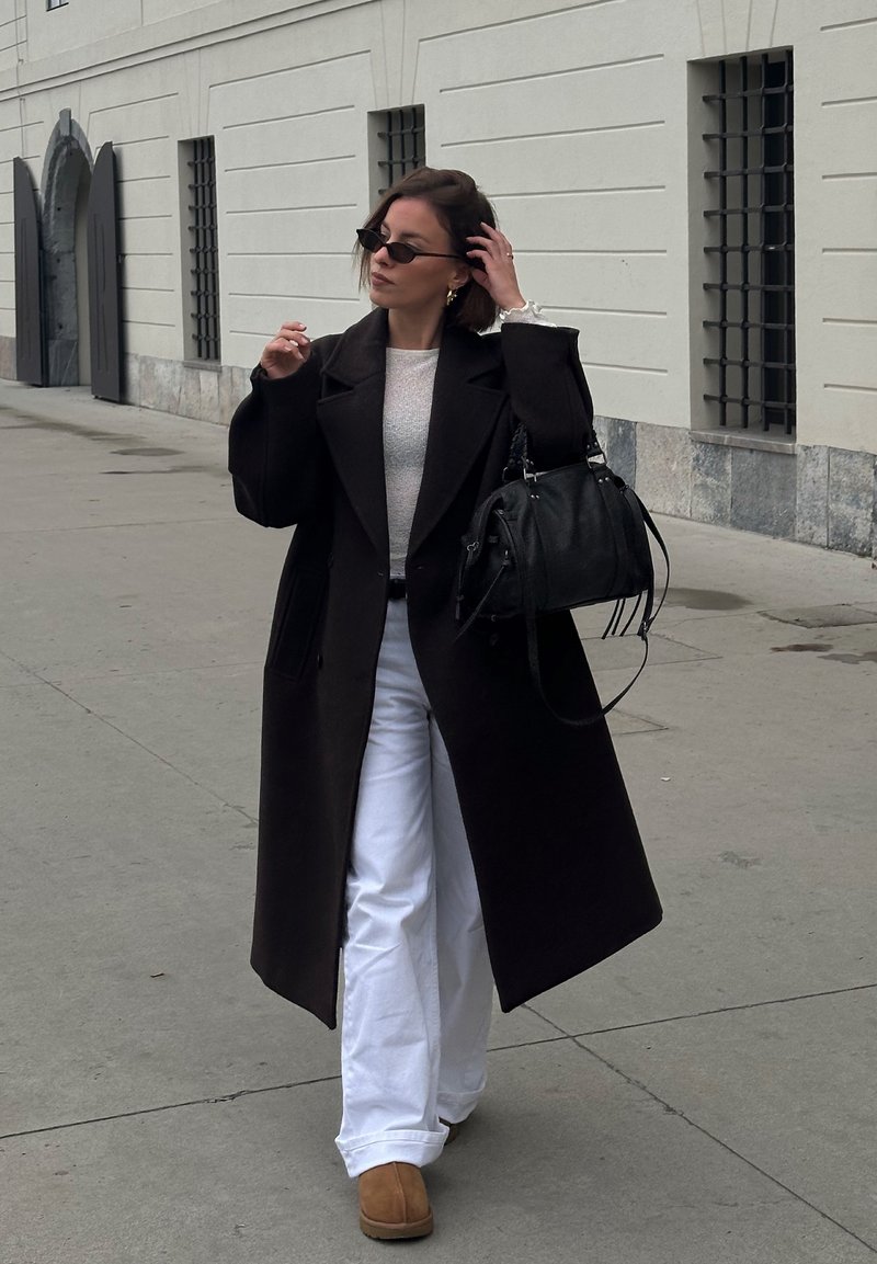 Palton lung din lână neagră peste un pulover deschis la culoare, pantaloni albi evazați și cizme maro. Geantă neagră cu curele. Cadru urban în aer liber.
