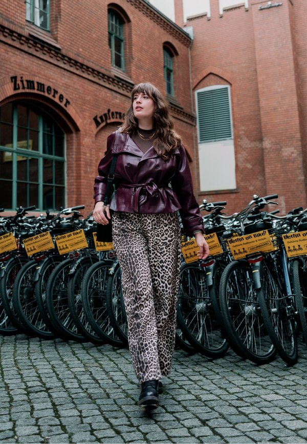 Femme portant une veste en cuir violette et un pantalon imprimé léopard marchant sur des pavés près d'une rangée de vélos de location avec des panneaux "Berlin on Bike".