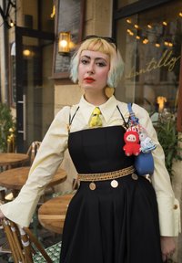 Persona con cabello rubio con puntas de colores, vistiendo una camisa amarilla, un vestido negro, accesorios dorados y llevando un bolso azul con llaveros de peluche.