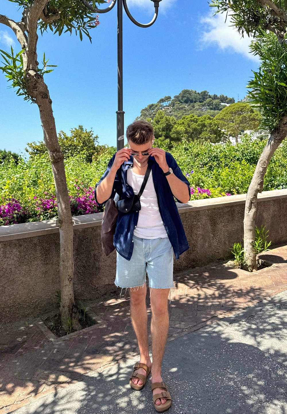 Short-sleeved navy shirt over a white tank top, denim shorts, brown sandals, and a crossbody bag. Surrounded by greenery and flowers.