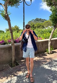 Short-sleeved navy shirt over a white tank top, denim shorts, brown sandals, and a crossbody bag. Surrounded by greenery and flowers.