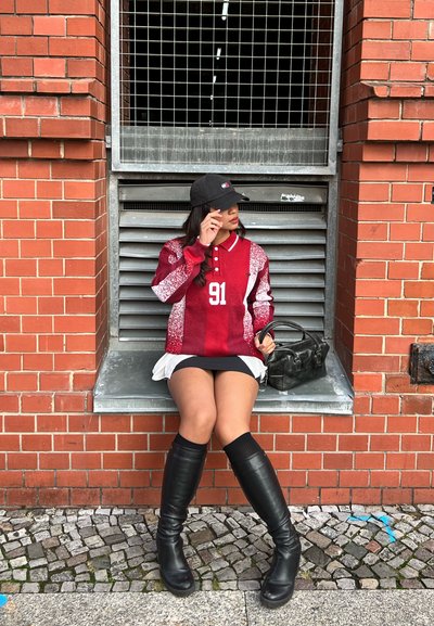 Camisa de manga larga a rayas rojas y blancas con el número 91, botas altas negras hasta la rodilla, gorra negra y un pequeño bolso negro. Fondo de muro de ladrillo.
