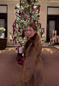 Kvinde i brun pelsjakke, der kigger tilbage nær et pyntet juletræ med røde bånd og indpakkede gaver indendørs.
