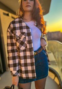 Hnědo-bílá kostkovaná košile s knoflíkovým zapínáním, nošená přes bílý top a džínovou sukni, doplněná hnědým páskem a černou kabelkou.