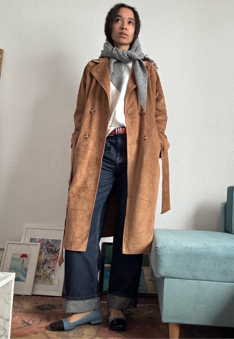 Une personne se tient à l'intérieur portant un trench en daim couleur fauve, une chemise blanche, une écharpe grise, un jean large et foncé, et des chaussures plates bicolores à petit talon.
